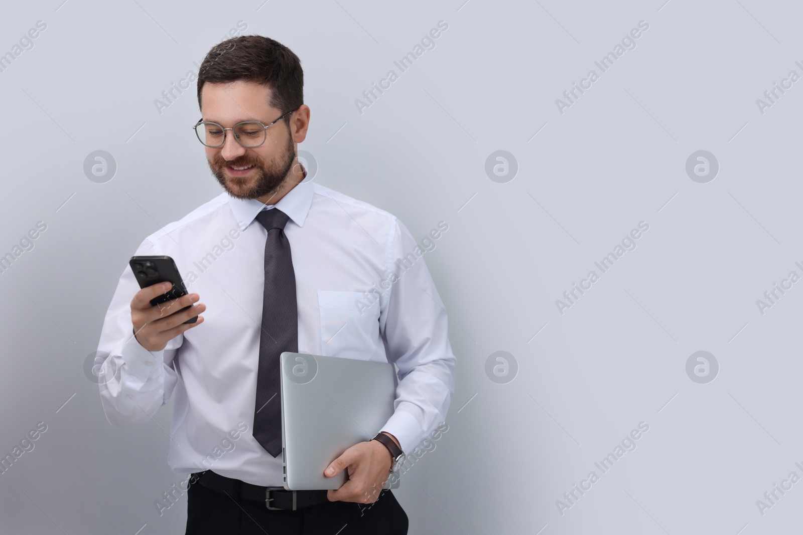 Photo of Portrait of smiling businessman with laptop using smartphone on grey background. Space for text