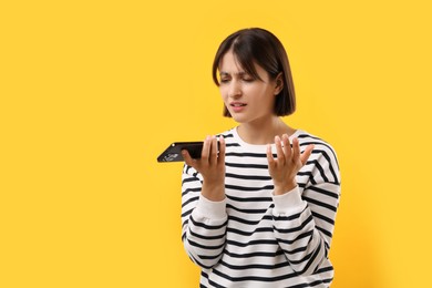 Unhappy woman recording voice message via smartphone on yellow background, space for text