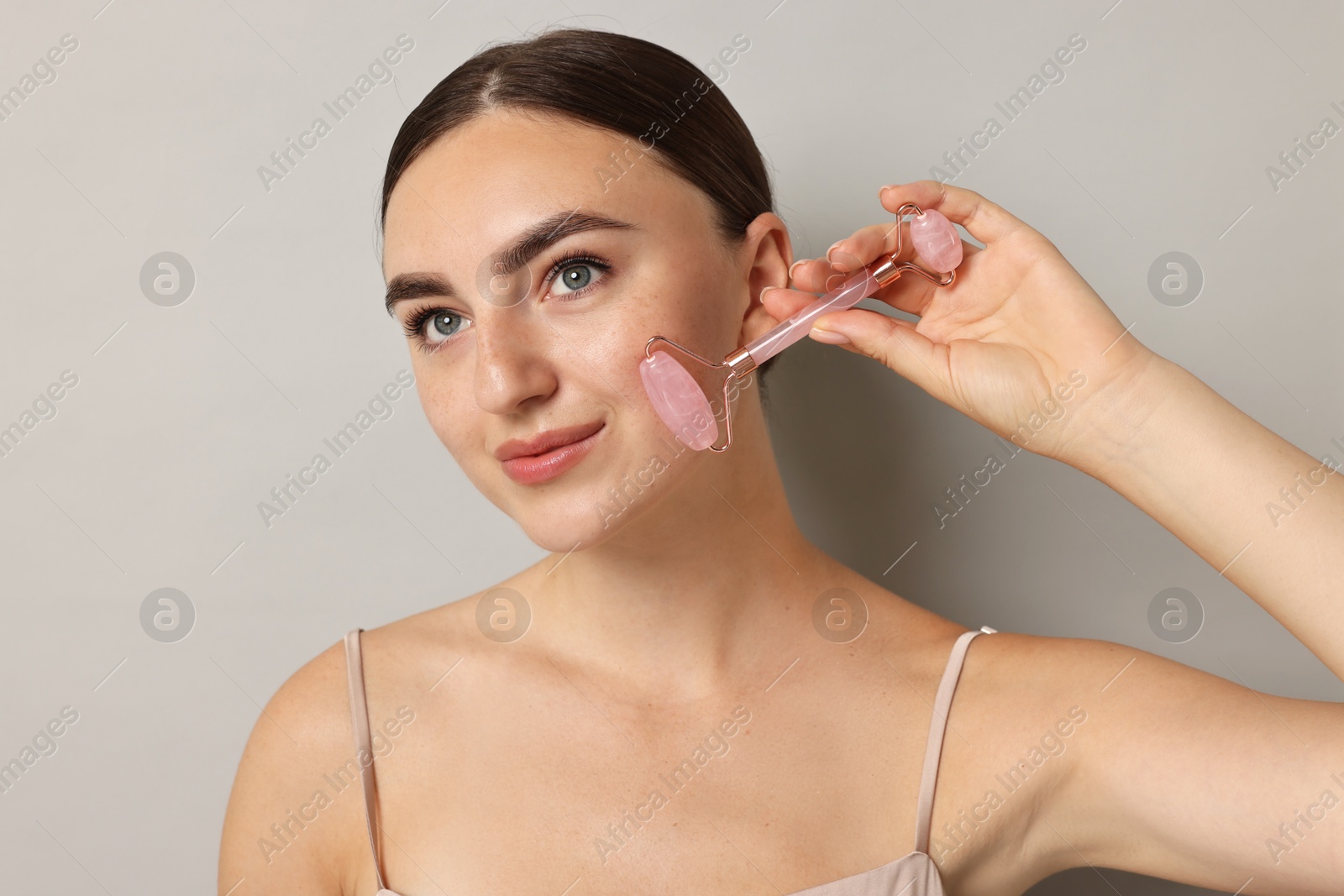 Photo of Beautiful young woman doing facial massage with roller on grey background