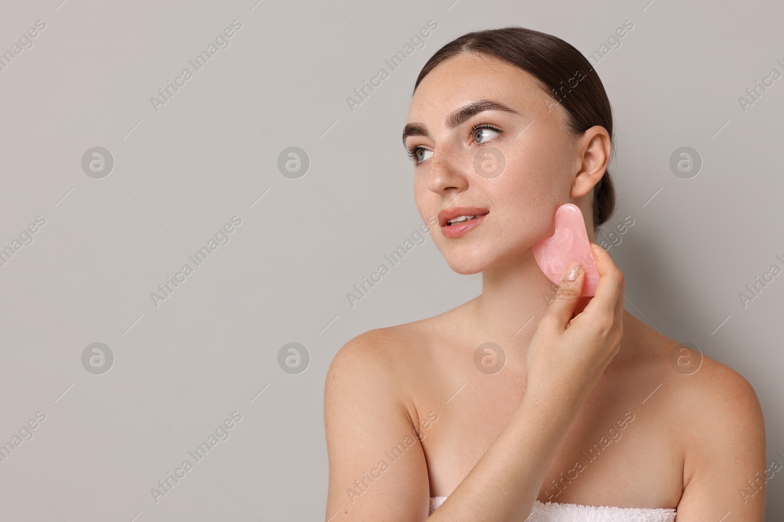 Photo of Beautiful young woman doing facial massage with gua sha tool on grey background, space for text