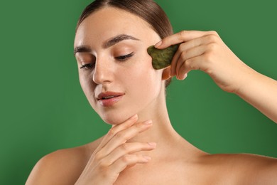 Photo of Beautiful young woman doing facial massage with gua sha tool on green background