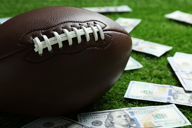 Photo of American football ball and dollar banknotes on green grass outdoors, closeup