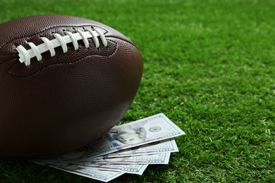 Photo of American football ball and dollar banknotes on green grass outdoors, space for text