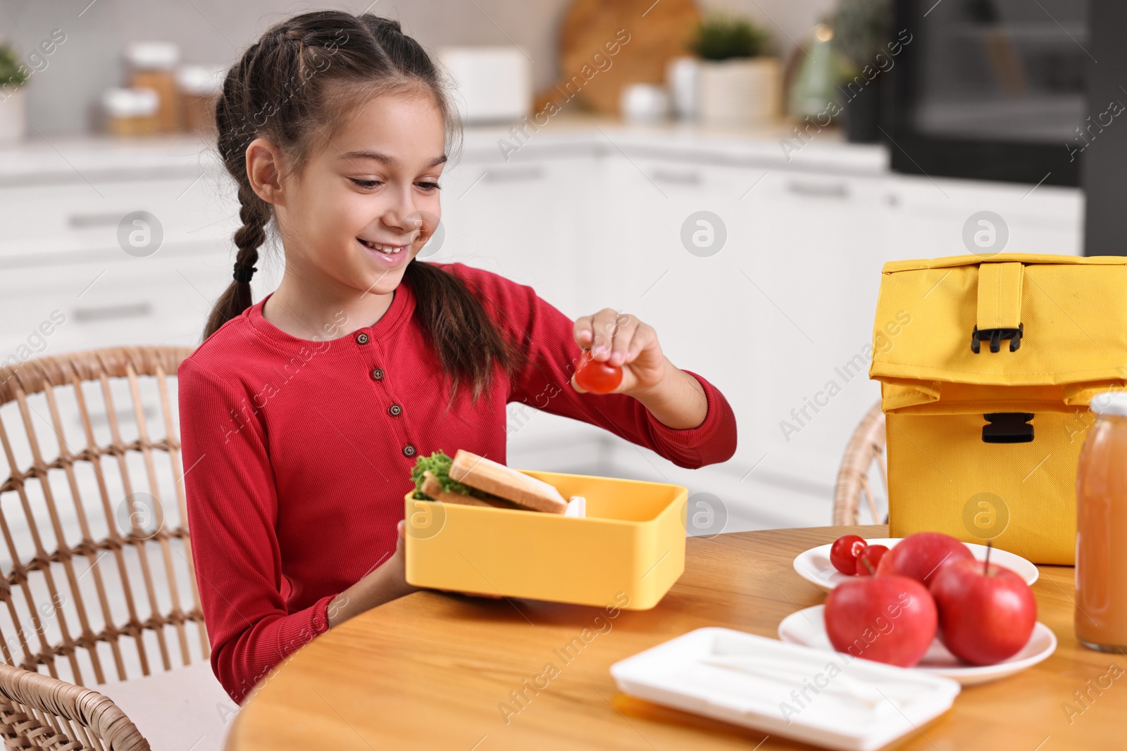 Photo of Cute girl putting tomato into lunch box at wooden table in kitchen. Preparing for school