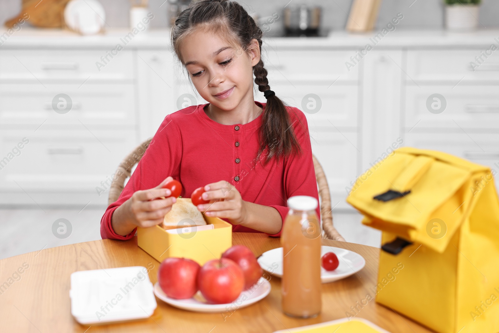 Photo of Cute girl putting tomatoes into lunch box at wooden table in kitchen. Preparing for school