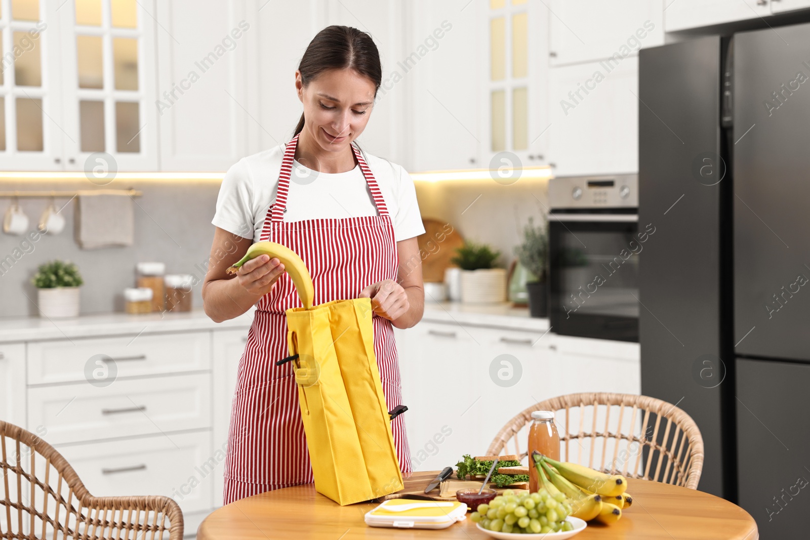 Photo of Woman putting fresh banana into lunch bag at wooden table in kitchen