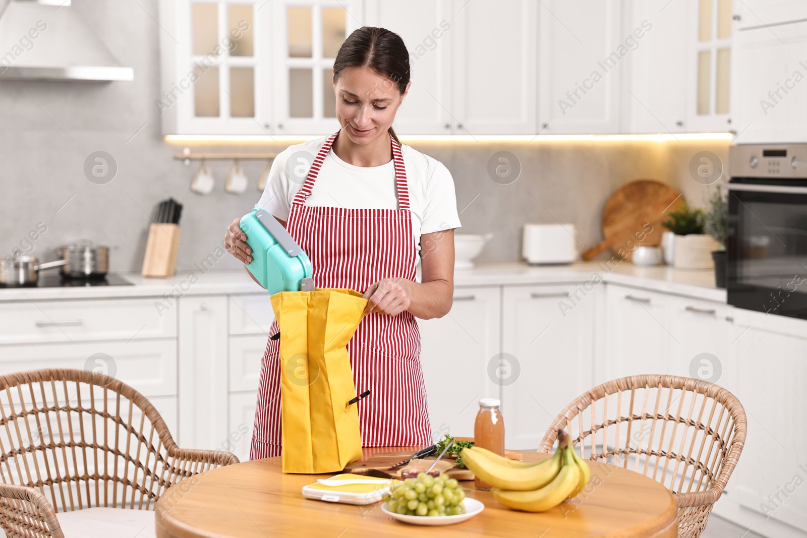 Photo of Woman putting school lunch box into bag at wooden table in kitchen