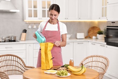 Woman putting school lunch box into bag at wooden table in kitchen