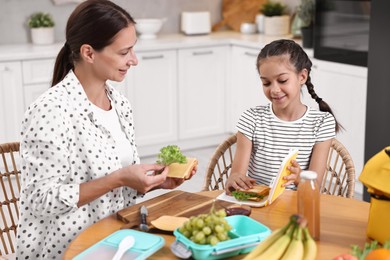 Cute girl and her mother packing lunch box with healthy products at wooden table in kitchen