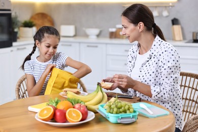 Cute girl and her mother preparing lunch box with healthy snacks at wooden table in kitchen