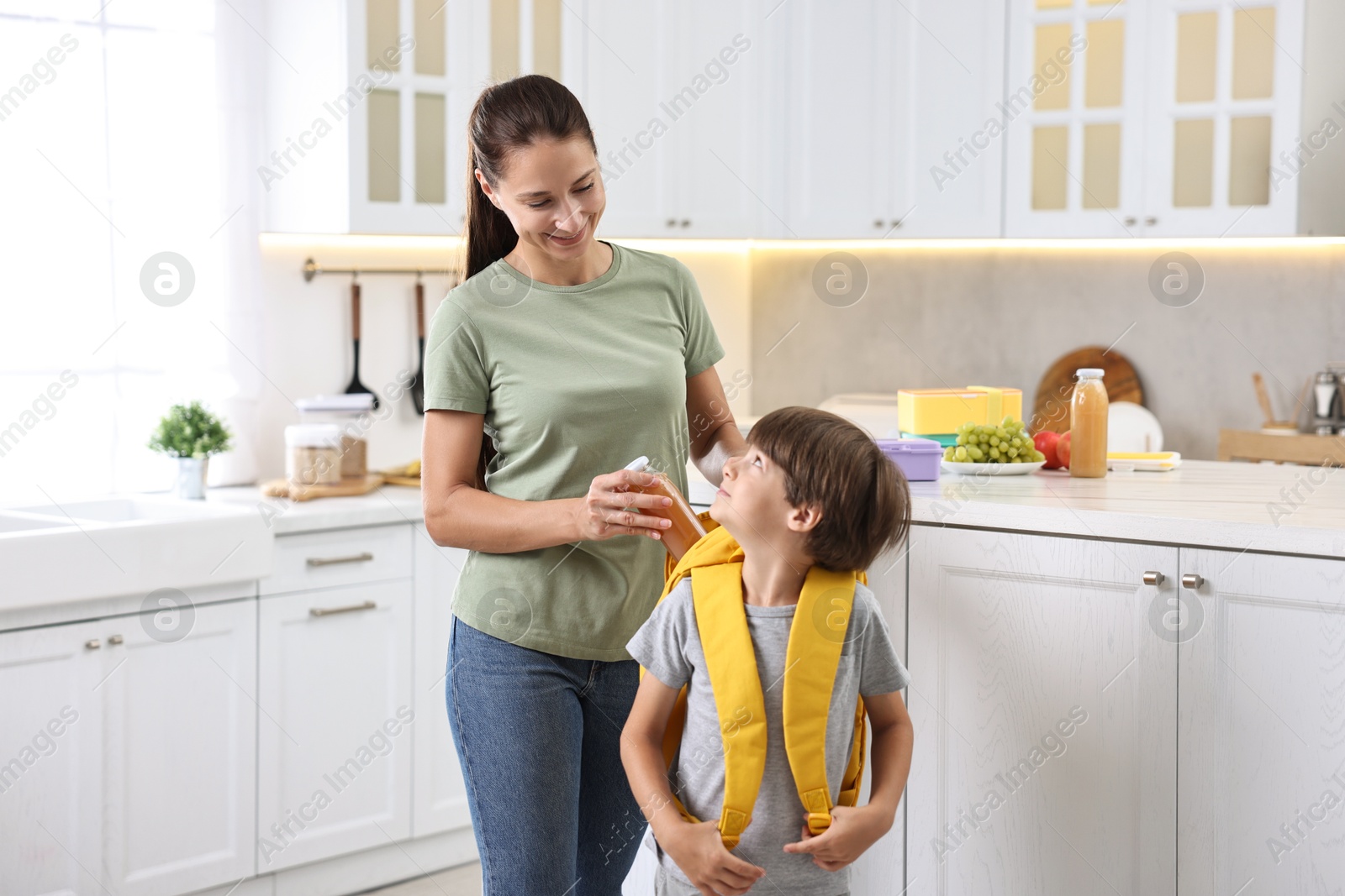 Photo of Smiling mother putting bottle of drink into son`s backpack in kitchen. Preparing for school