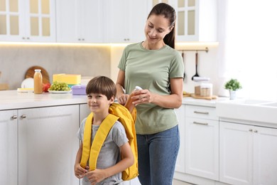 Smiling mother putting bottle of drink into son`s backpack in kitchen. Preparing for school