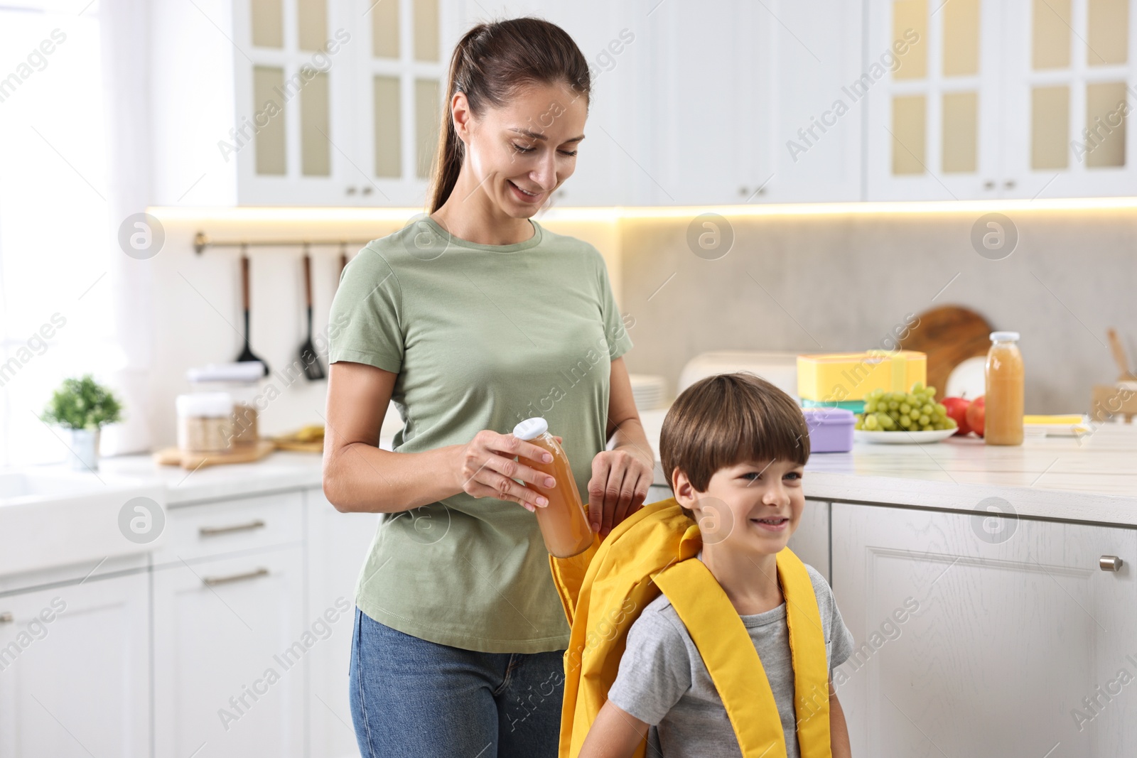 Photo of Smiling mother putting bottle of drink into son`s backpack in kitchen. Preparing for school