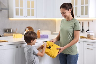 Smiling mother giving lunch bag to her son in kitchen