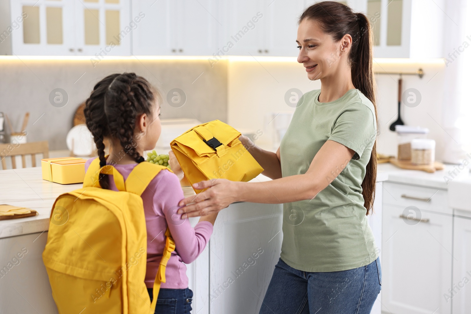 Photo of Smiling mother giving lunch bag to her daughter in kitchen. Preparing for school