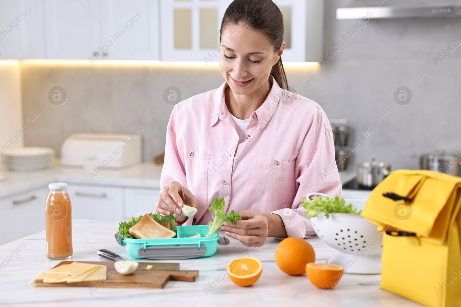 Photo of Smiling woman making snacks for school lunch box at white marble table in kitchen