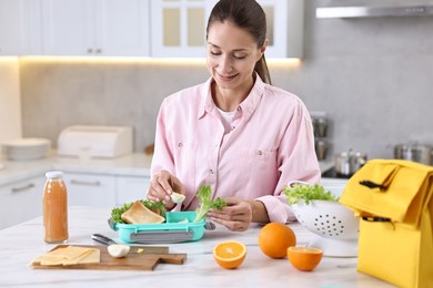 Smiling woman making snacks for school lunch box at white marble table in kitchen