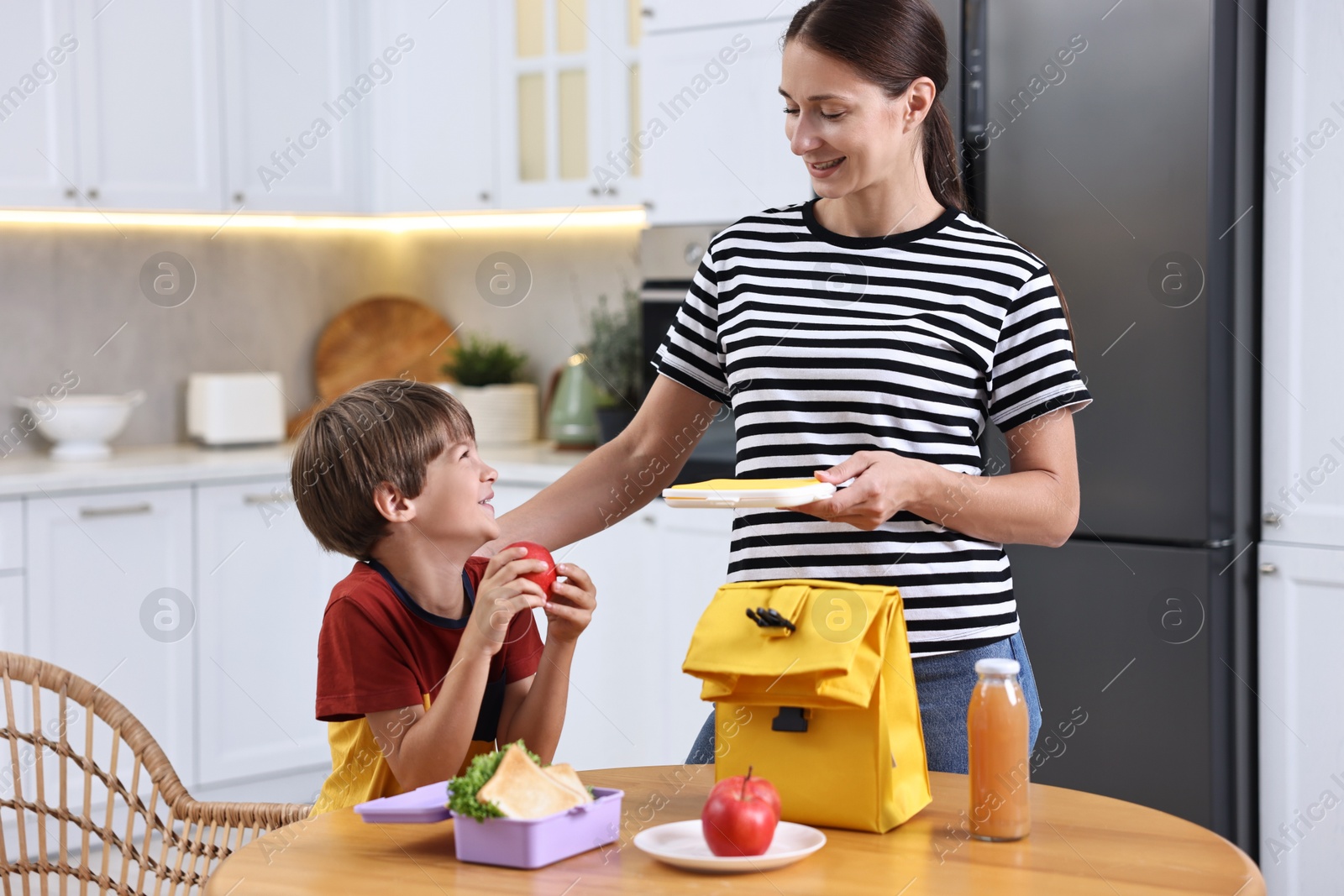 Photo of Smiling mother and her cute son preparing school lunch box with healthy food at wooden table in kitchen