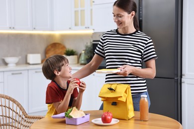 Photo of Smiling mother and her cute son preparing school lunch box with healthy food at wooden table in kitchen