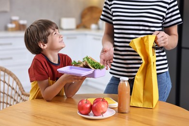 Photo of Mother and her cute son preparing school lunch box with healthy food at wooden table in kitchen, closeup