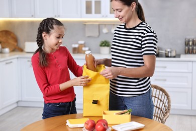 Photo of Mother with her daughter putting bottle of drink into lunch bag at wooden table in kitchen. Preparing for school