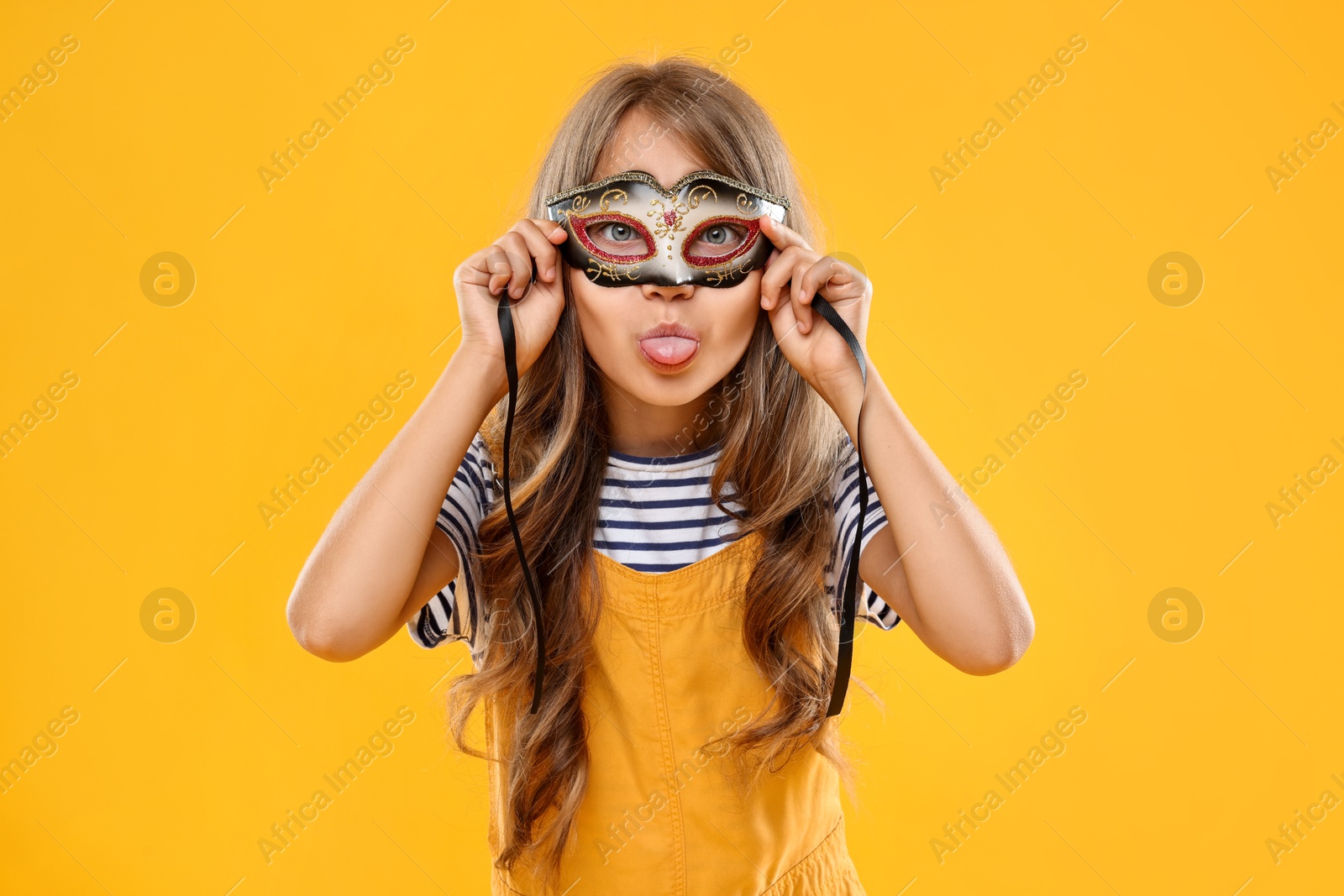 Photo of Cute girl wearing carnival mask and showing tongue on orange background