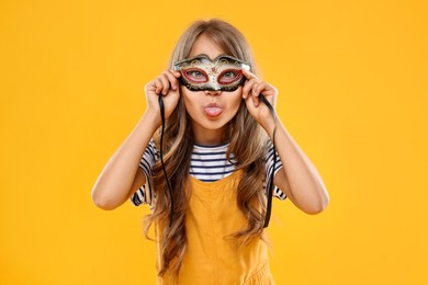 Photo of Cute girl wearing carnival mask and showing tongue on orange background