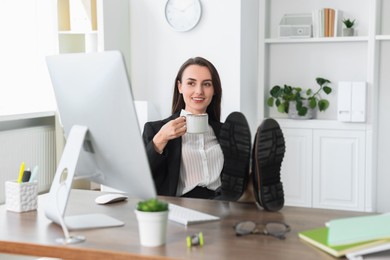 Smiling businesswoman drinking coffee holding legs on table in office. Break time