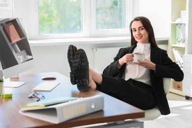 Photo of Smiling businesswoman drinking coffee holding legs on table in office. Break time