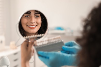 Photo of Patient looking in mirror while doctor holding teeth color chart in clinic, closeup. Dental veneers