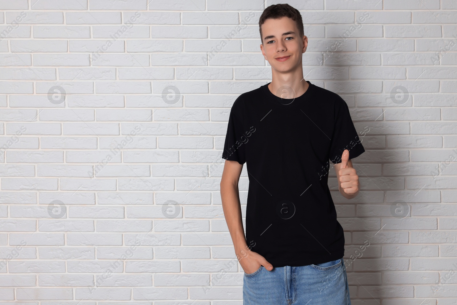 Photo of Teenage boy wearing black t-shirt and showing thumbs up near white brick wall, space for text