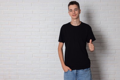 Teenage boy wearing black t-shirt and showing thumbs up near white brick wall, space for text