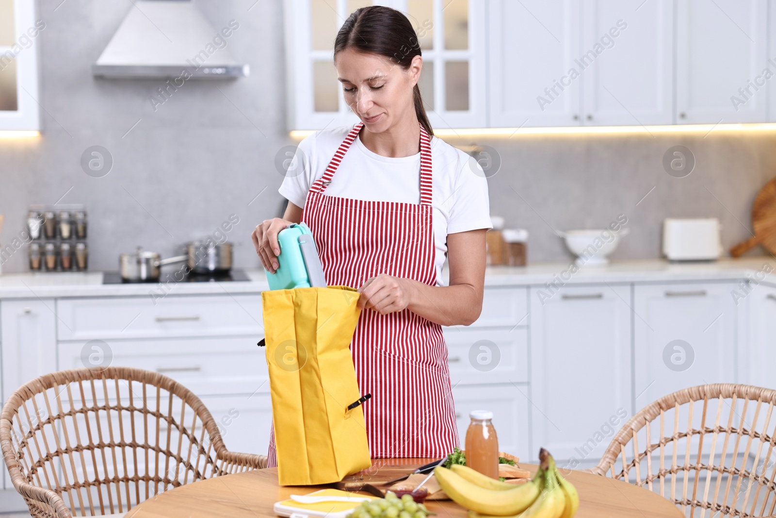 Photo of Woman putting school lunch box into bag at wooden table in kitchen