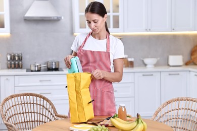 Woman putting school lunch box into bag at wooden table in kitchen