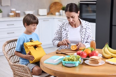 Mother and her cute son preparing lunch box with healthy food at wooden table in kitchen