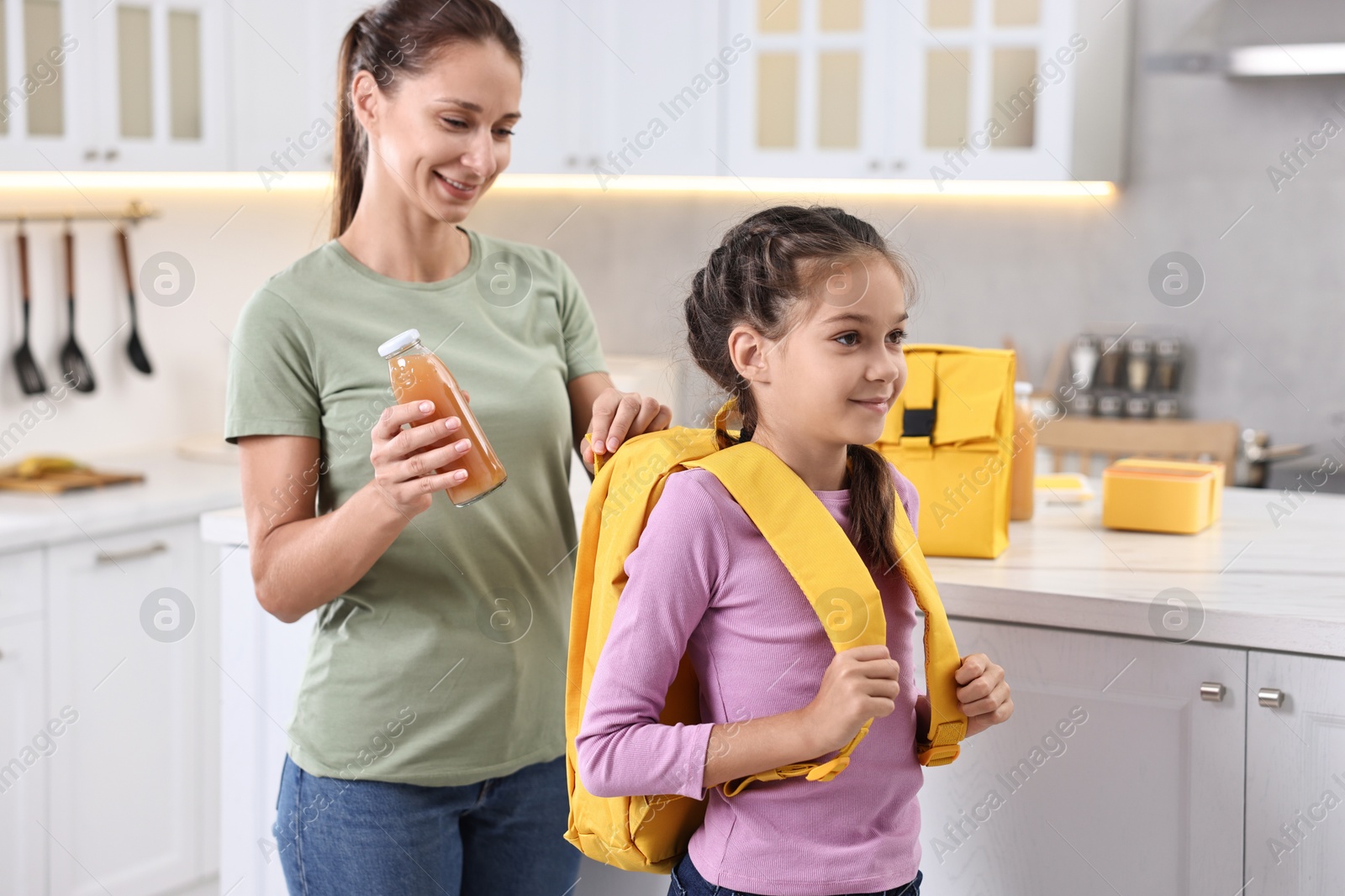 Photo of Smiling mother putting bottle of drink into daughter`s backpack in kitchen