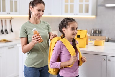 Smiling mother putting bottle of drink into daughter`s backpack in kitchen