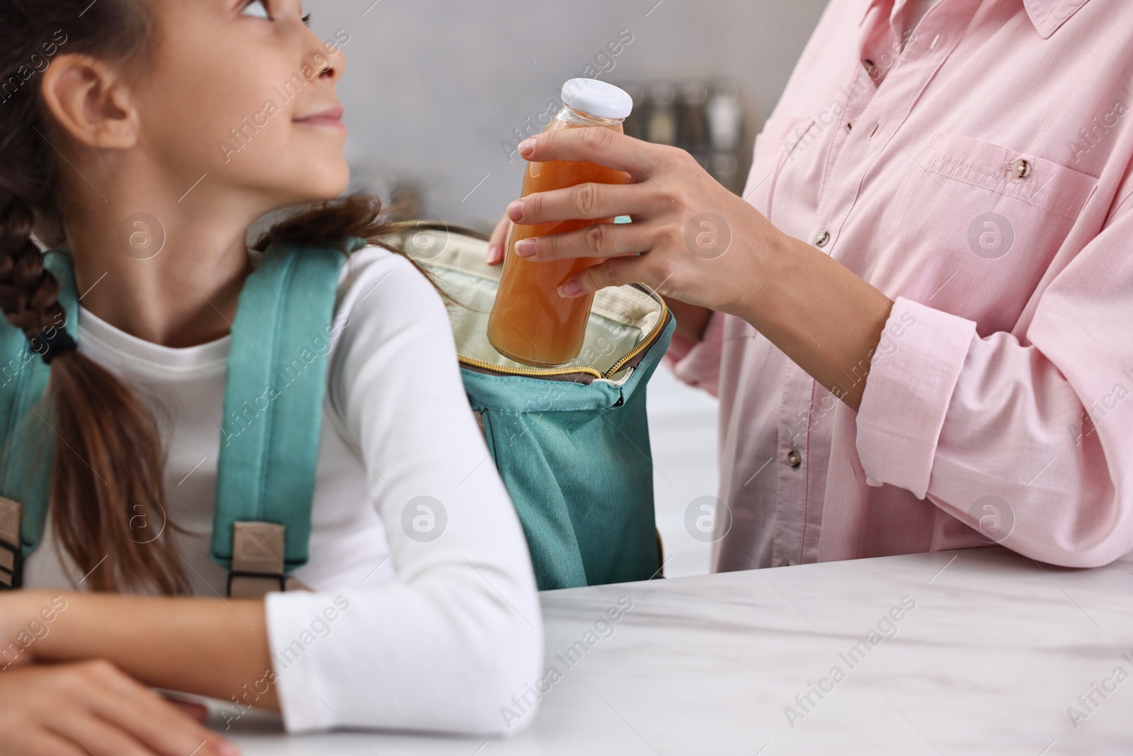 Photo of Mother putting bottle of drink into daughter`s backpack in kitchen, closeup. Preparing for school