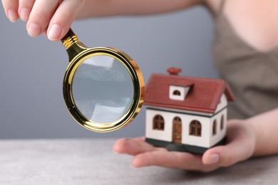 House hunting. Woman with magnifying glass and house model at grey table, closeup