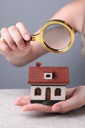 House hunting. Woman with magnifying glass and house model at grey table, closeup