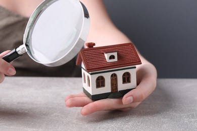 House hunting. Woman with magnifying glass and house model at grey table, closeup