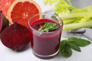 Photo of Tasty beetroot smoothie with basil in glass, fresh vegetables and grapefruits on light tiled table, closeup. Vegan drink