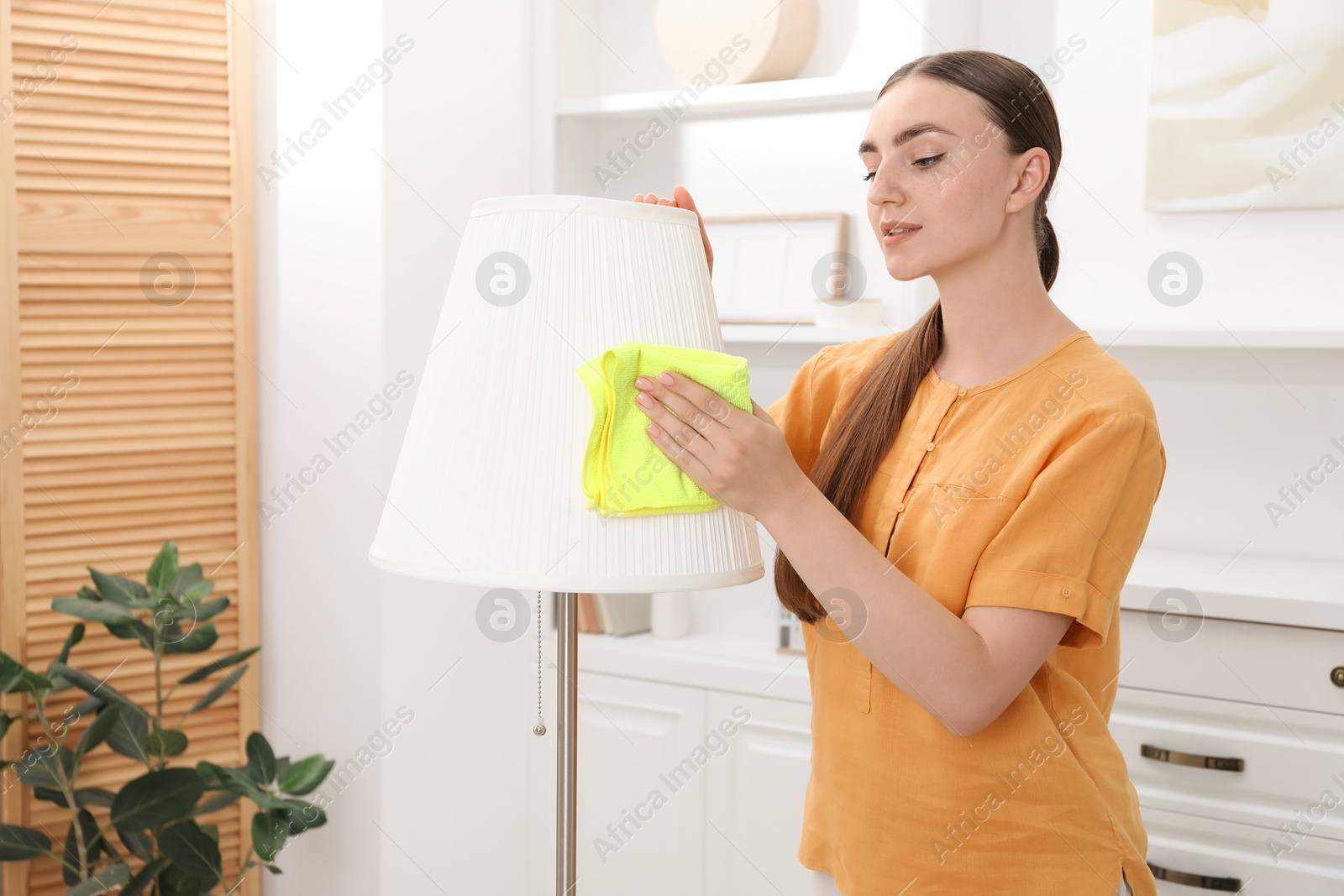 Photo of Beautiful young woman cleaning floor lamp with rag at home