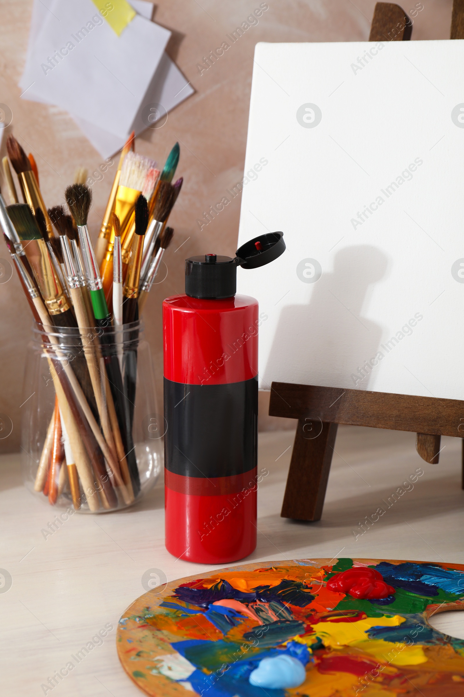 Photo of Red acrylic paint in bottle, small easel, palette and brushes on wooden table indoors