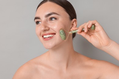 Beautiful young woman doing facial massage with roller on grey background