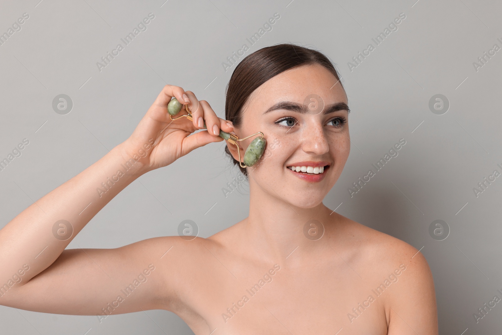 Photo of Beautiful young woman doing facial massage with roller on grey background