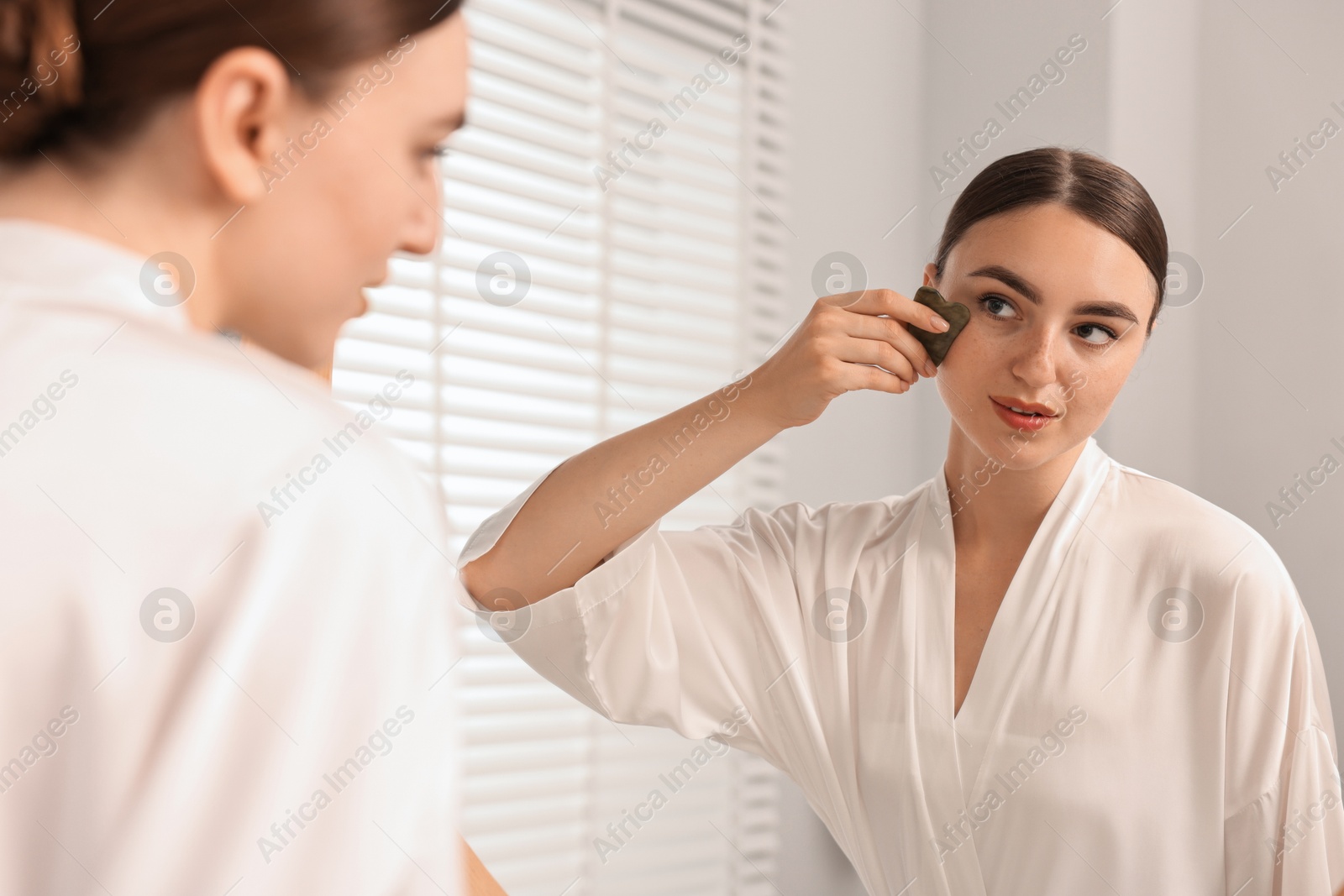 Photo of Beautiful young woman doing facial massage with gua sha tool near mirror at home