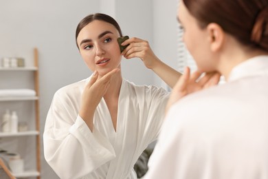 Photo of Beautiful young woman doing facial massage with gua sha tool near mirror at home