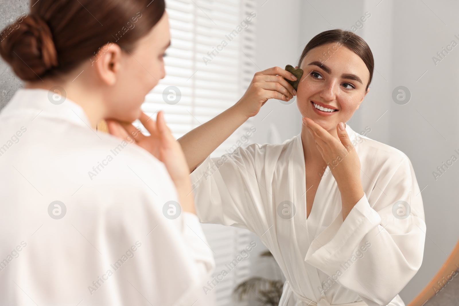 Photo of Beautiful young woman doing facial massage with gua sha tool near mirror at home