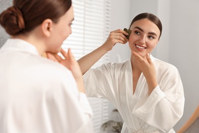 Beautiful young woman doing facial massage with gua sha tool near mirror at home